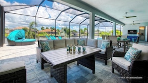 Een overdekt terras met rieten meubilair, een tafel met flessen en uitzicht op een woonwijk met huizen en palmbomen.