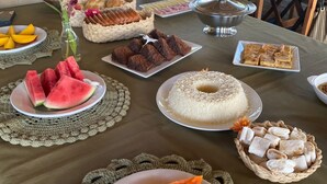A table with various food items, including watermelon, bread, and a cake, set on a tablecloth with a crocheted doily. - Pousada Quintal da Praia  (Prado)