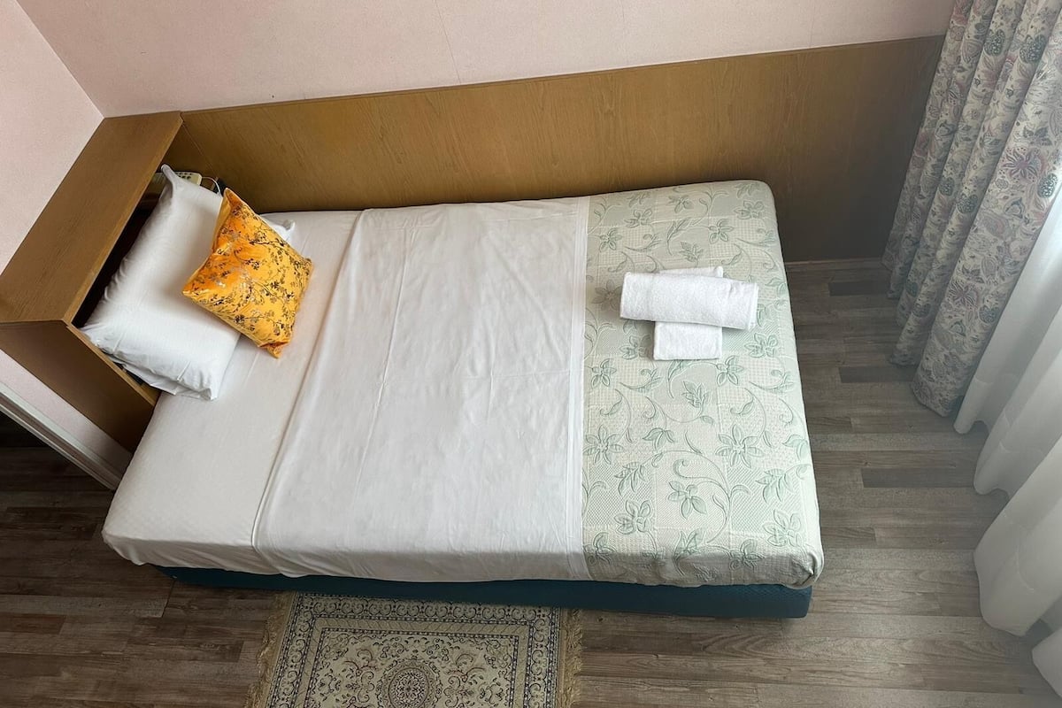 A small, neatly made bed with white and patterned bedding, a yellow pillow, and two white towels.
