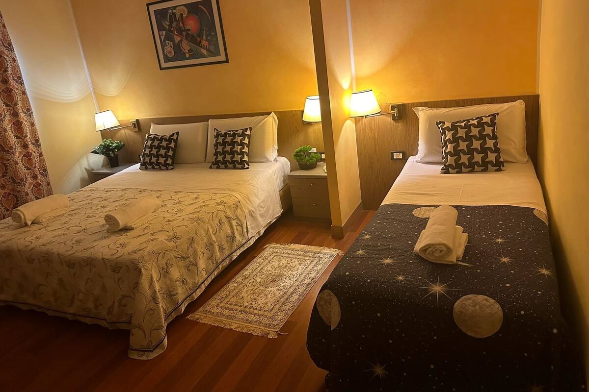 A hotel room with two beds, each with patterned bedding and pillows, a nightstand with a lamp, a small potted plant, and a framed picture on the wall.
