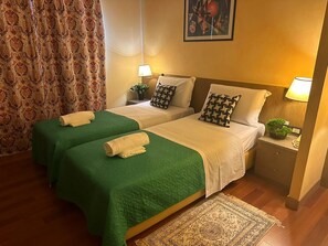 Ein Hotelzimmer mit zwei Betten, grünen Bettdecken, weißen Laken, gemusterten Kissen, Nachttischlampen, einem kleinen Topfpflanzen und einem dekorativen Teppich.