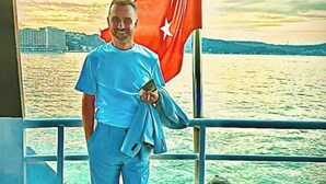 Eine Person steht auf dem Deck eines Bootes, eine Flagge im Arm, mit Jacke und sichtbarer Reling.