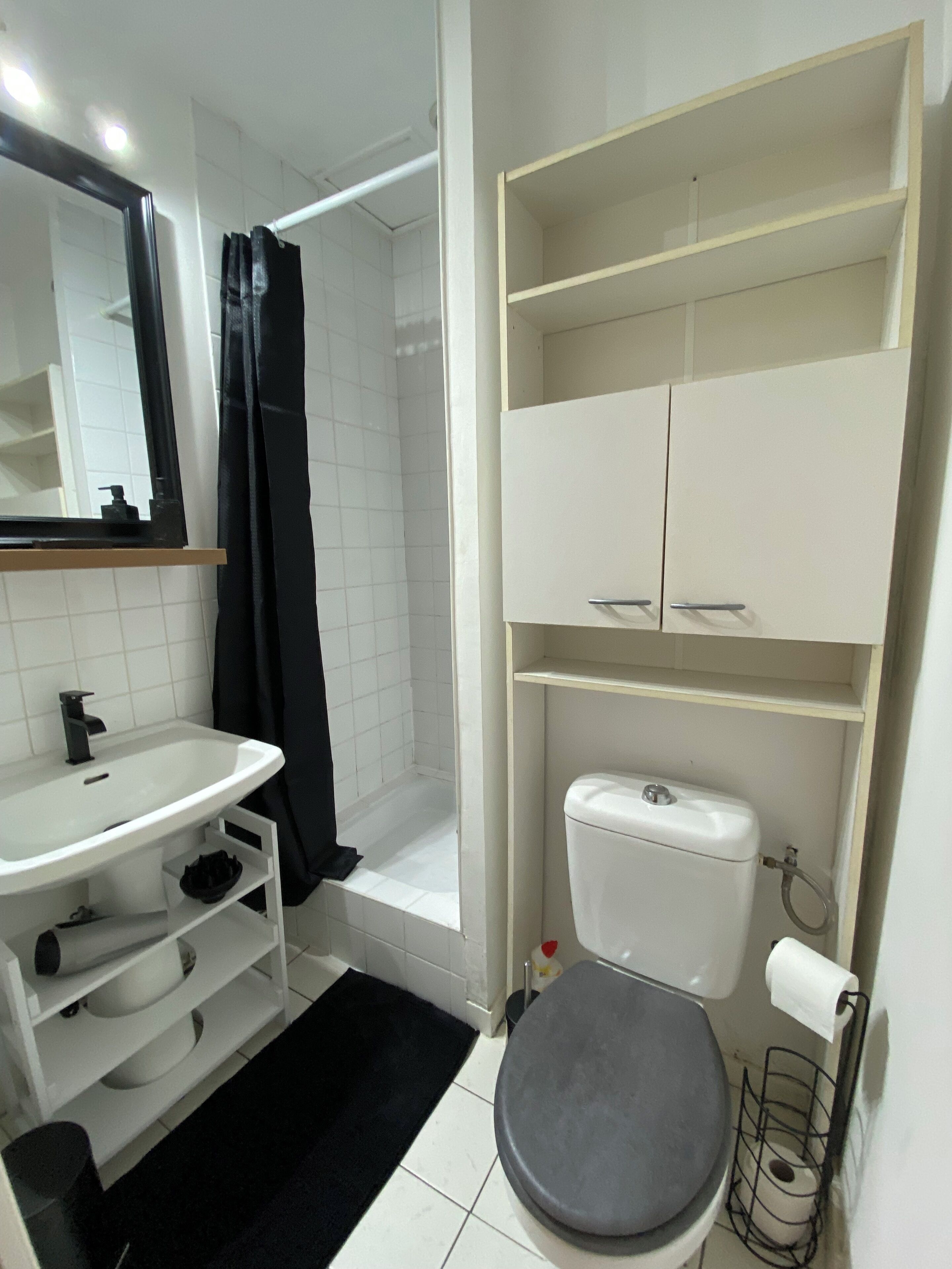 Une salle de bain avec un lavabo, des toilettes et une douche, le tout de couleur blanche. Il y a des étagères au-dessus des toilettes et un miroir au-dessus du lavabo.