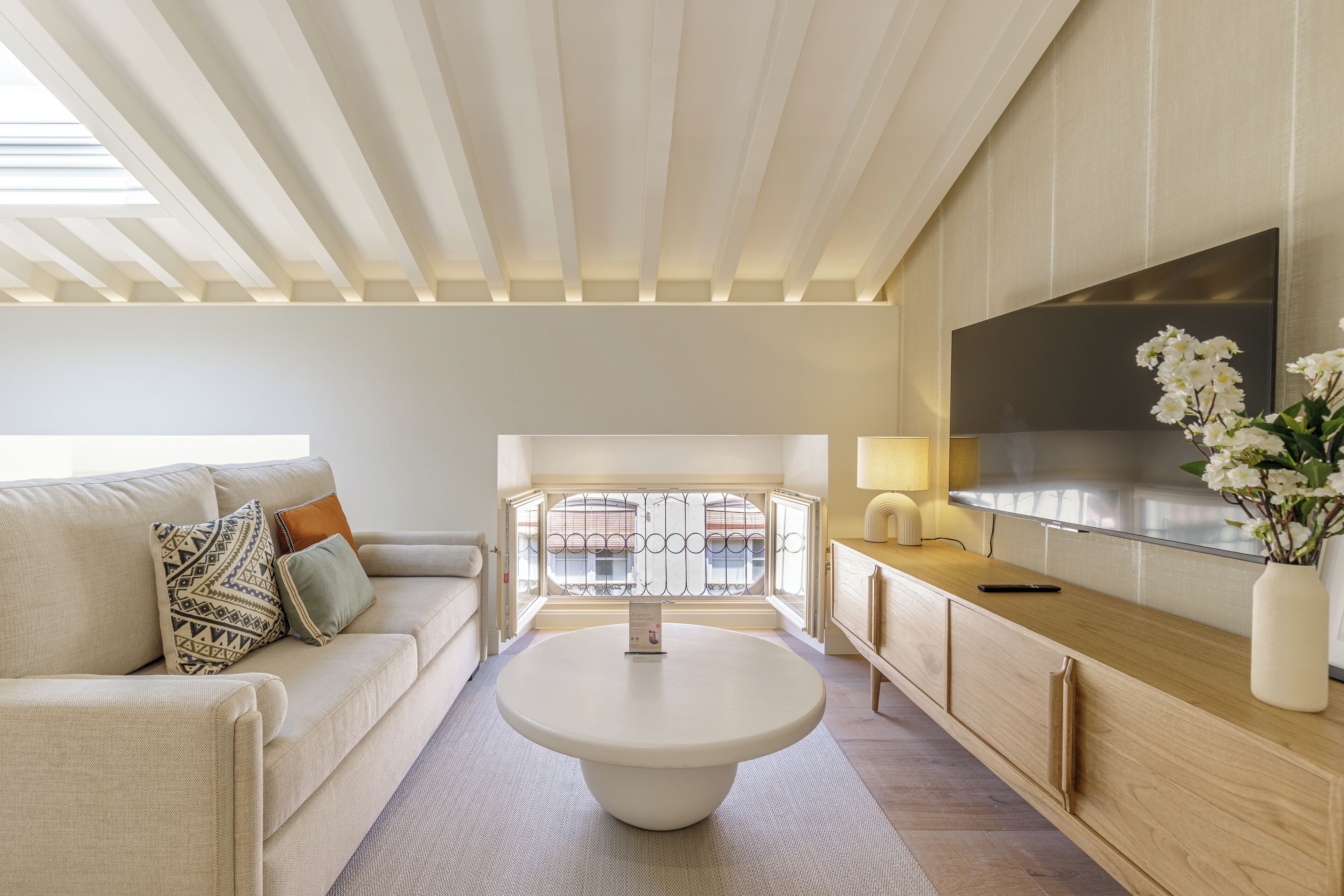 A modern living room with a round coffee table, a sofa with patterned cushions, a TV mounted on the wall, and a view through a window with a balcony.