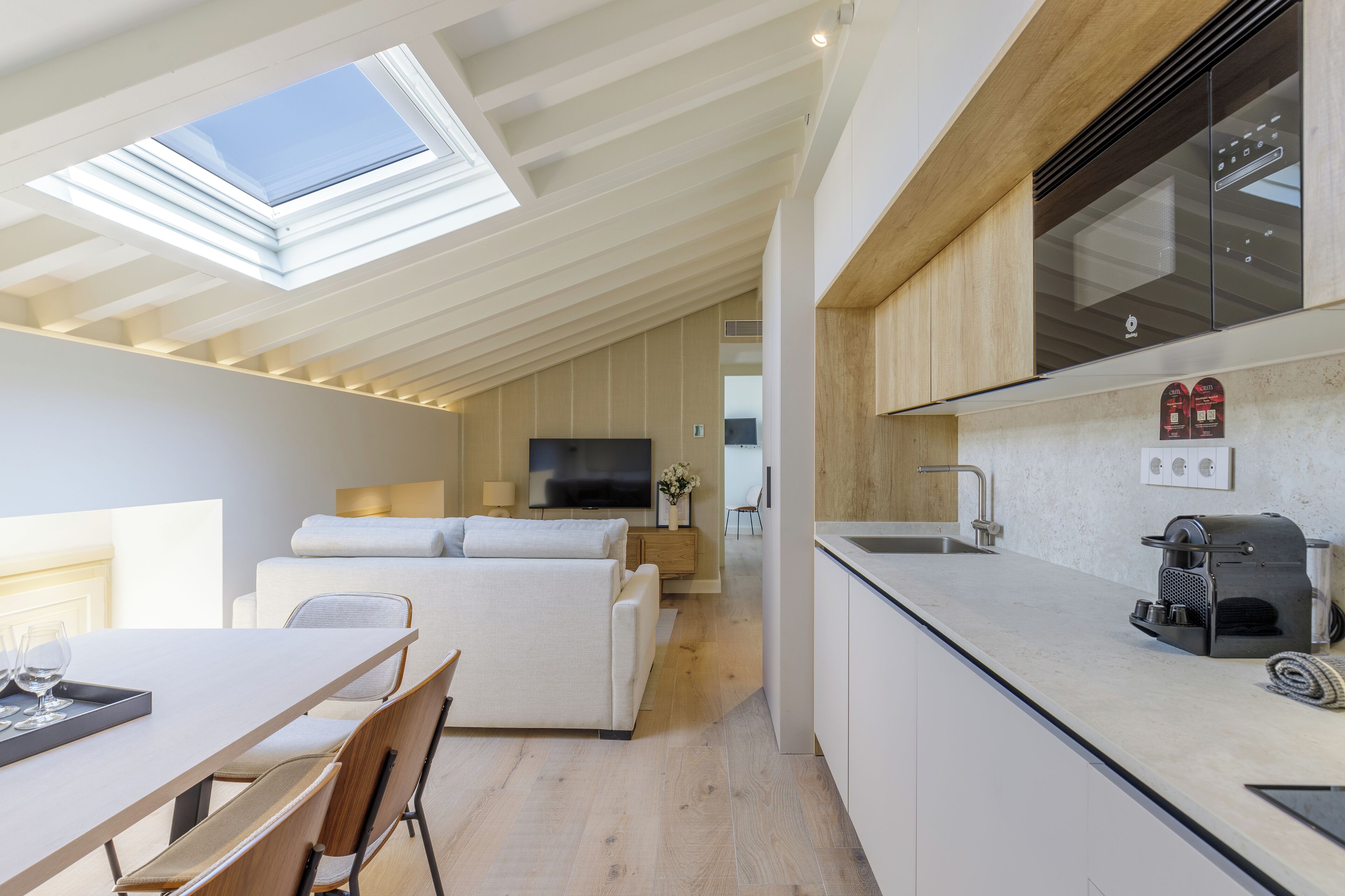A modern kitchen with a built-in oven and a coffee machine, a dining area with a table and chairs, and a living area with a sofa and a TV.