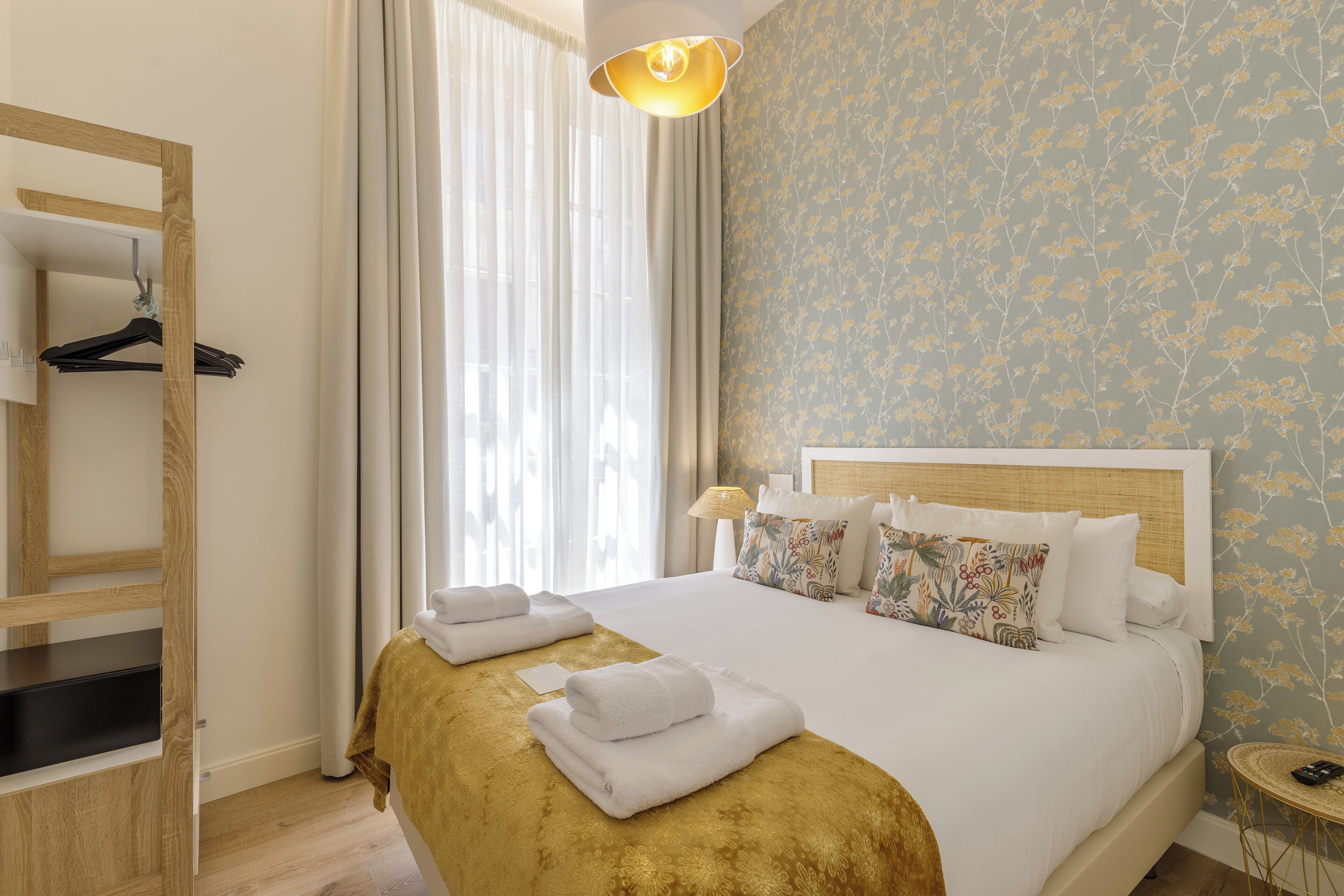 Een hotelkamer met een bed, een houten kledingkast, een spiegel, een raam met gordijnen en een wand met bloemenbehang.