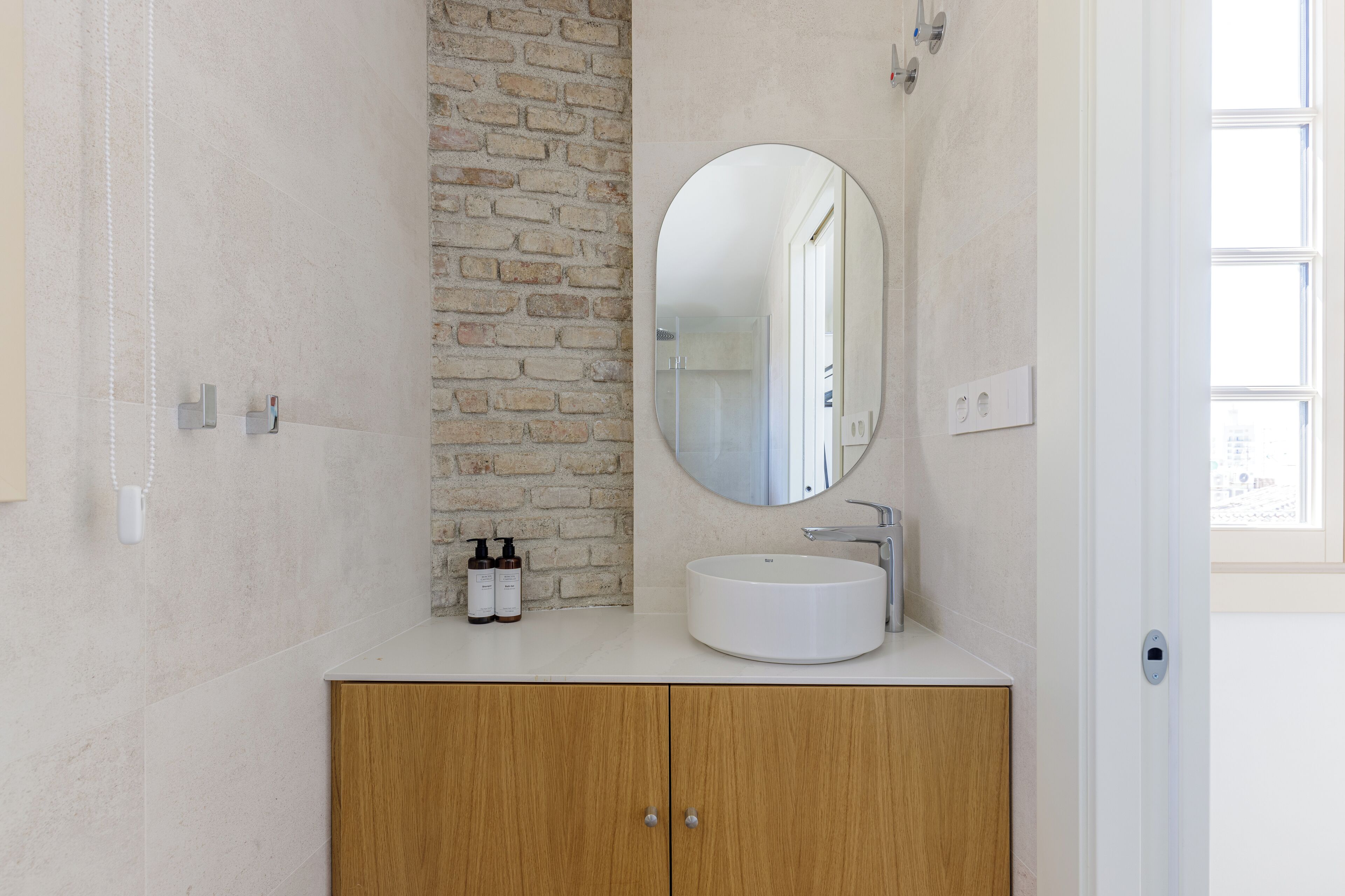Un baño con un espejo redondo, un lavabo blanco, armarios de madera y una pared de ladrillo decorativa.
