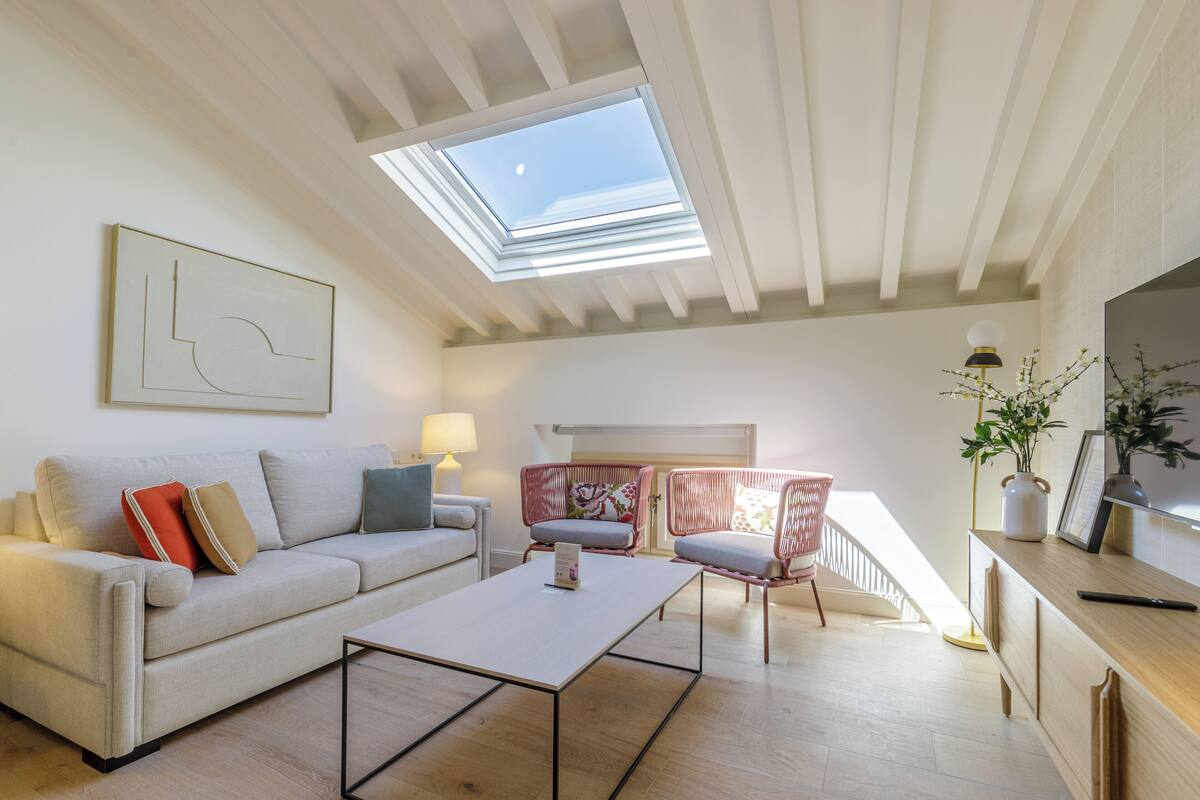 Sala de estar con sofá, mesa de centro y dos sillones. Hay un tragaluz sobre el sofá y una escalera que sube a un nivel superior.