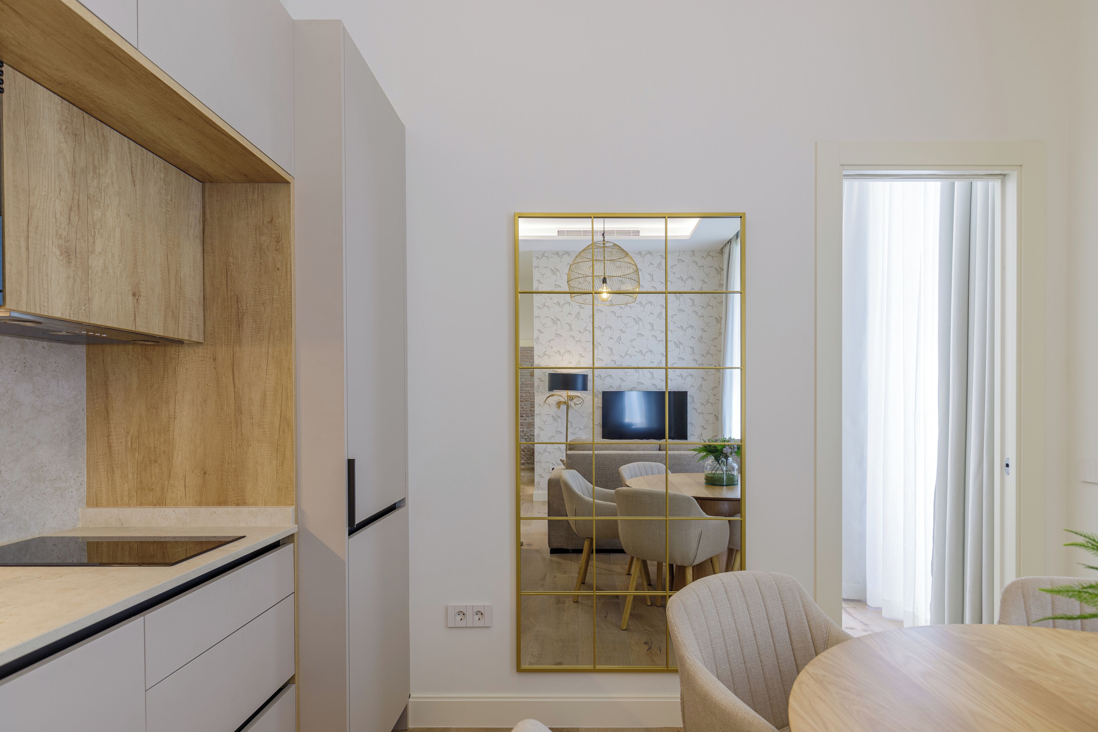 Una cocina moderna con armarios de madera, una zona de comedor con una mesa redonda y sillas, y un espejo grande con diseño de cuadrícula.