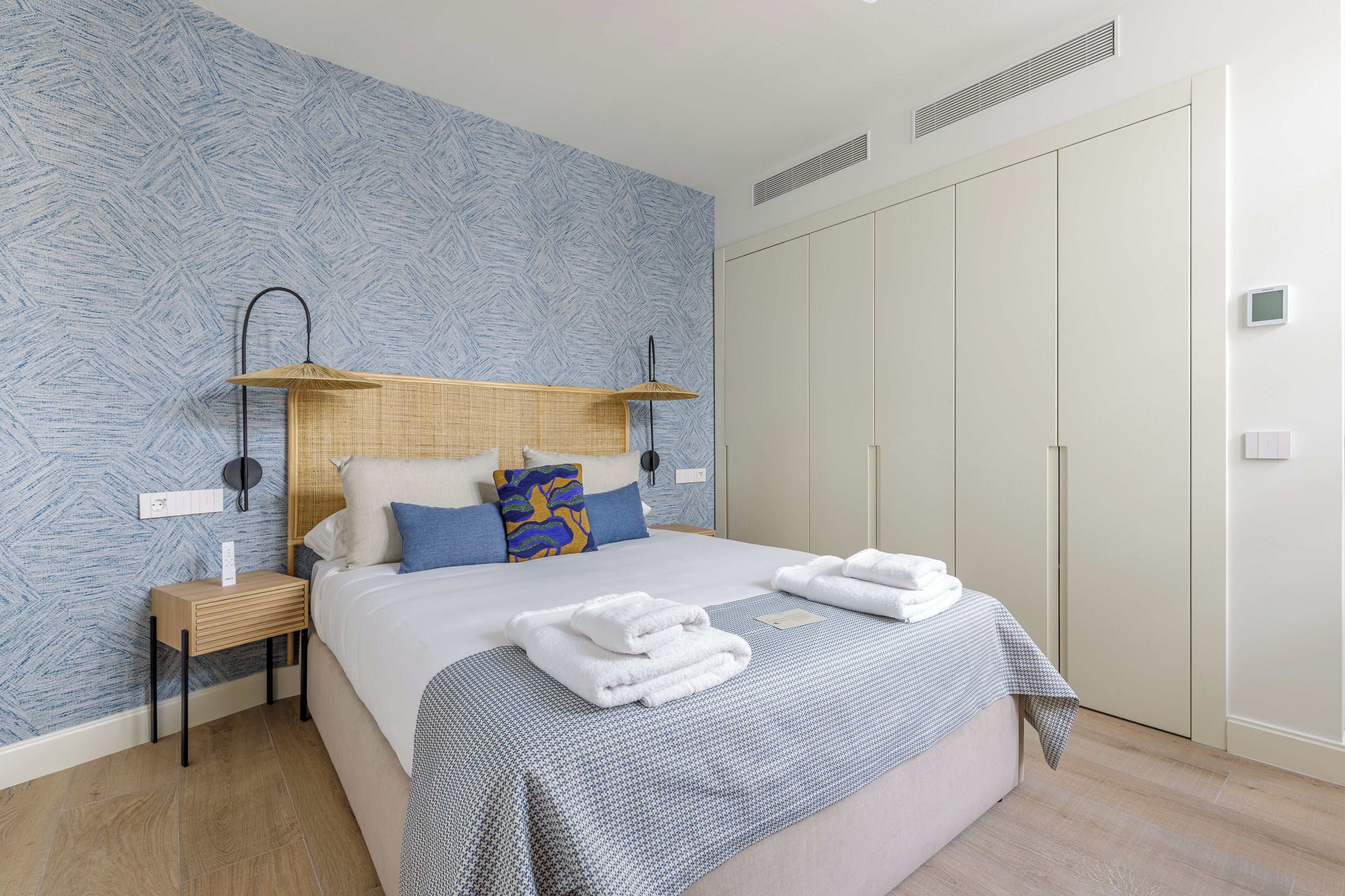 Ein ordentlich gemachtes Bett mit weißen Leinen und einer karierten Decke, ein Nachttisch mit Lampe sowie ein Kleiderschrank mit Spiegel.