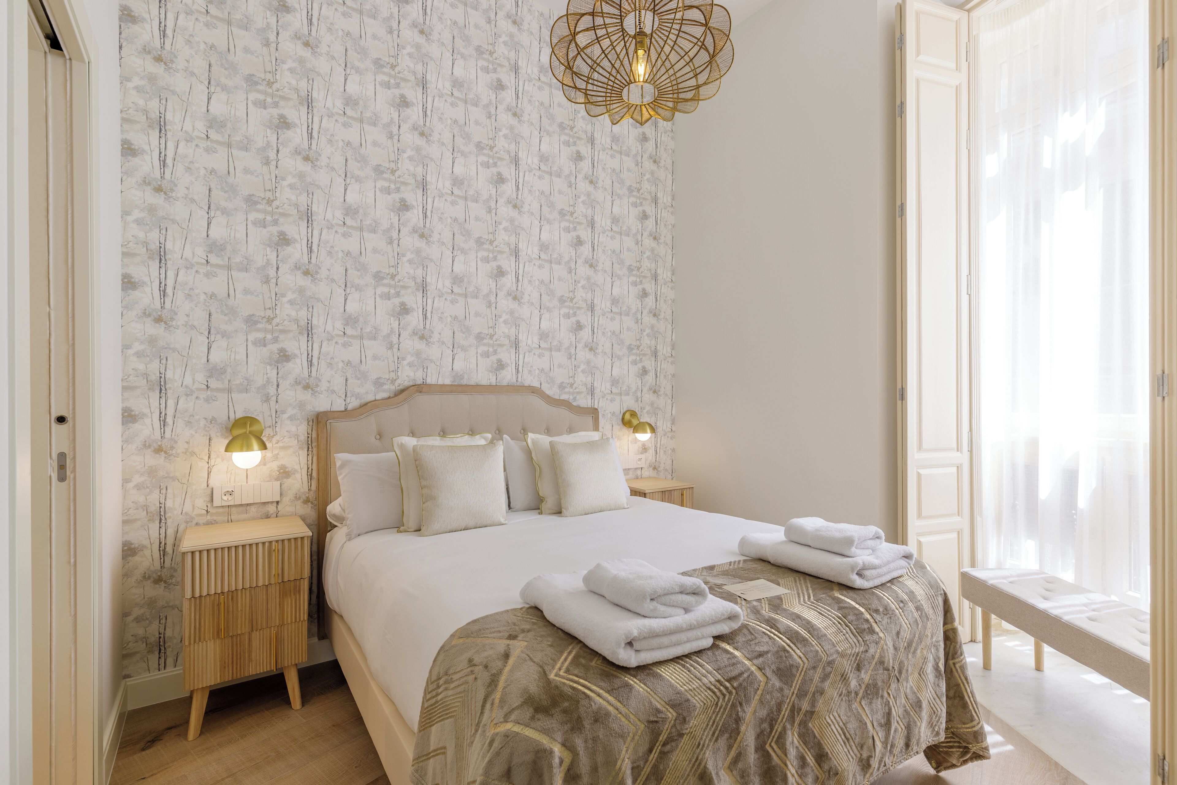 Un dormitorio con papel tapiz estampado, una cama con cubrecama estampado, dos lámparas de noche y una lámpara colgante.