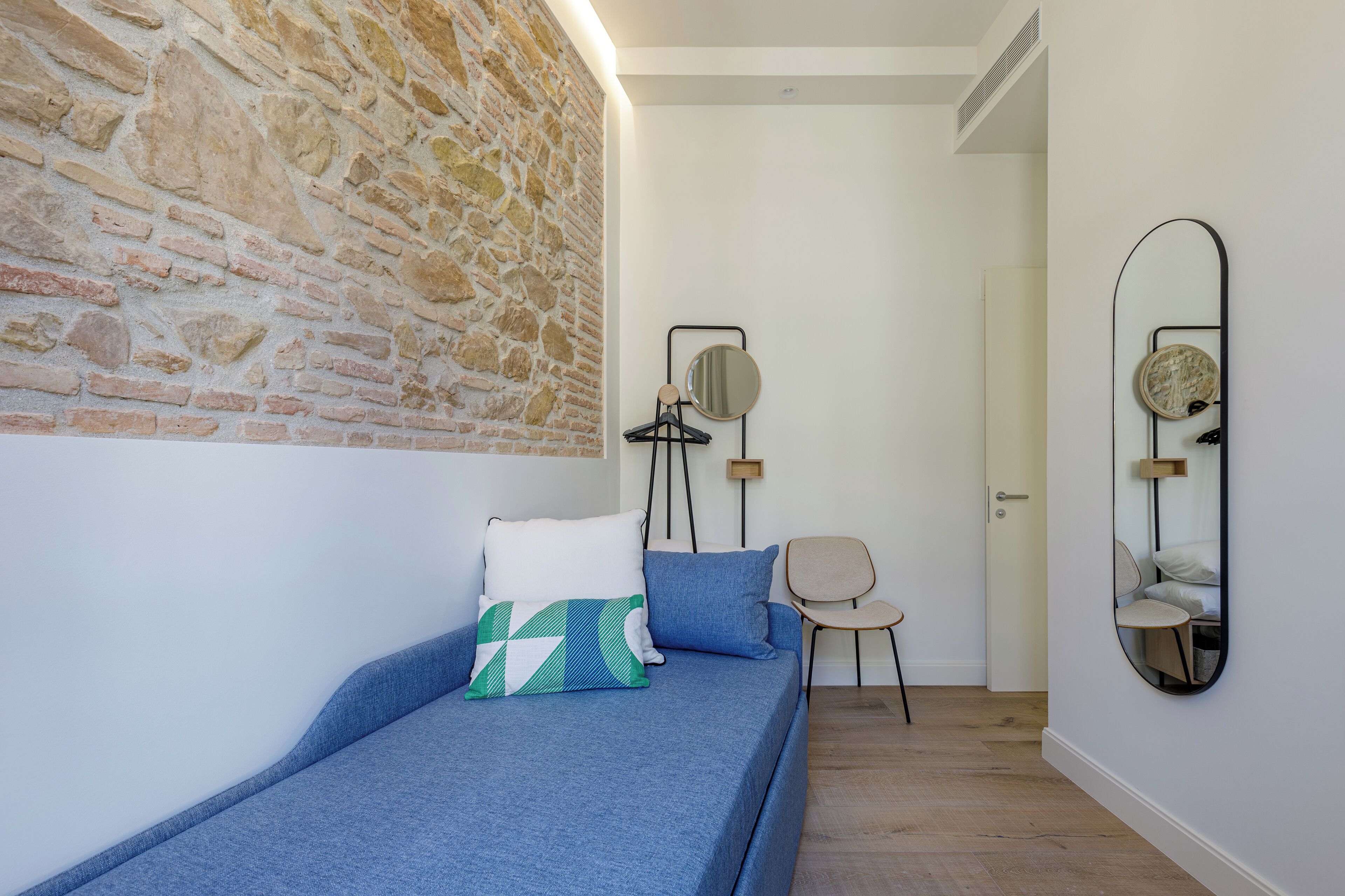 Een slaapkamer met een stenen muur, een bed met blauw beddengoed, een stoel en twee spiegels aan de muur.