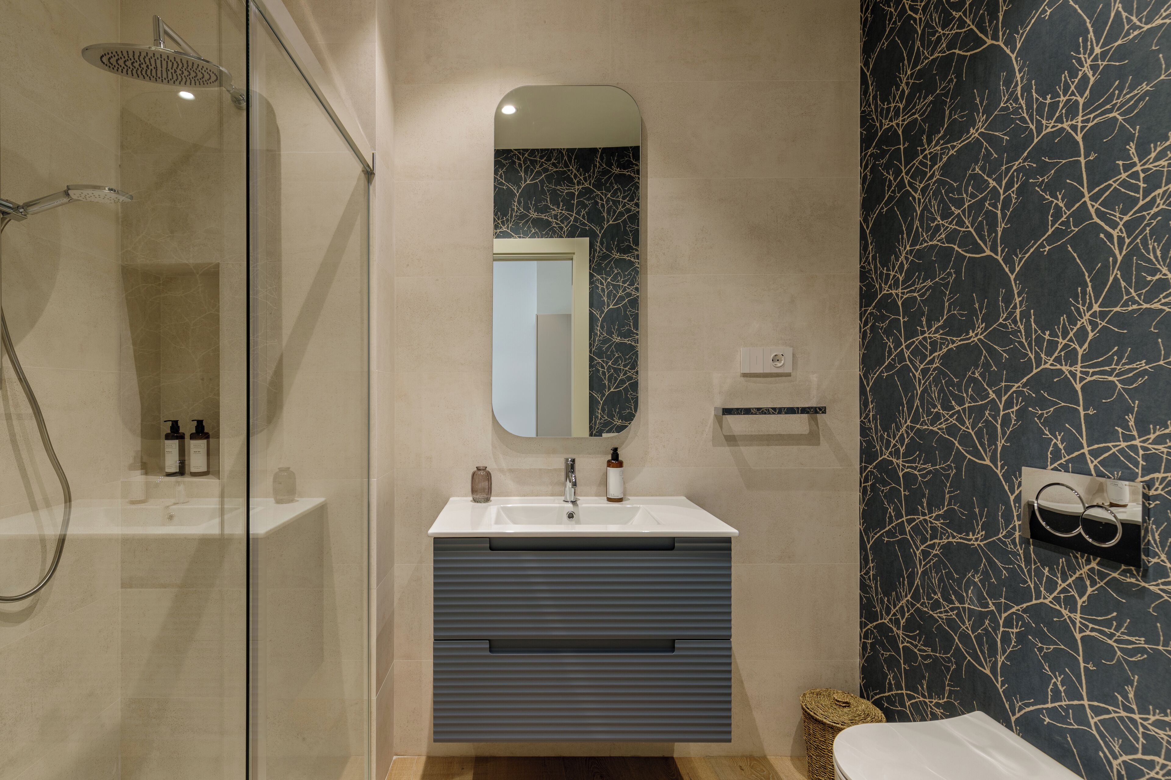 Ein modernes Badezimmer mit einer gläsernen Duschkabine, einem Waschtisch inklusive Spiegel und einer Wand mit Baumzweig-Muster.