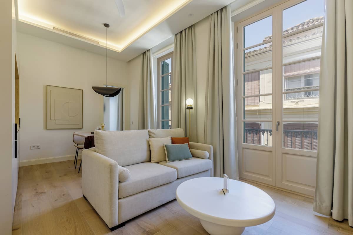 Una sala moderna con un sofá, una mesa redonda de centro, una lámpara colgante y vistas de un edificio a través de ventanas amplias.