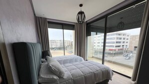 A modern bedroom with a large bed, a view of a cityscape through floor-to-ceiling windows, and a hanging light fixture. - Agadir Bay, a comfy apartment with pool (Agadir)