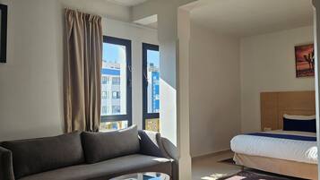 Quarto de hotel moderno com sofá, mesa redonda pequena, cama e janelas amplas com vista para a paisagem urbana.