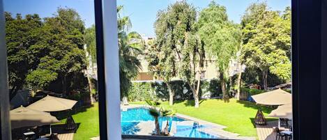 Vista desde una ventana a un patio trasero con piscina, muebles de jardín y abundante vegetación.