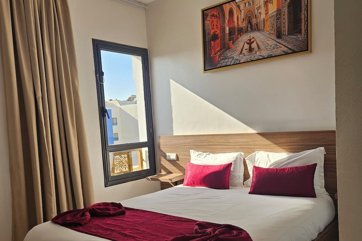 Une chambre d’hôtel avec un grand lit, une fenêtre avec des rideaux, un tableau au mur et un tapis à motifs.