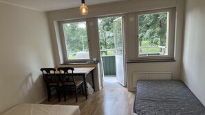 Une petite pièce lumineuse, dotée d’un plancher en bois, d’un lit, d’un bureau avec des chaises et de grandes fenêtres donnant sur un jardin.