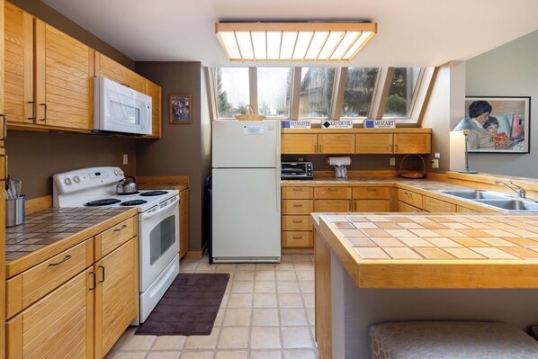 A kitchen with wooden cabinets, a microwave, stove, refrigerator, and a window with a view of trees.