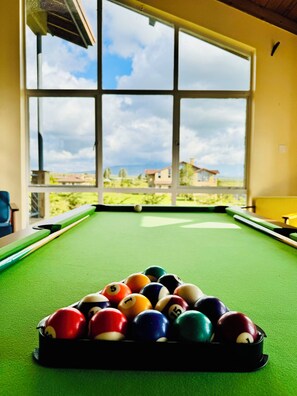 A pool table with balls arranged in a triangle, a window with a view of houses, and a yellow wall.