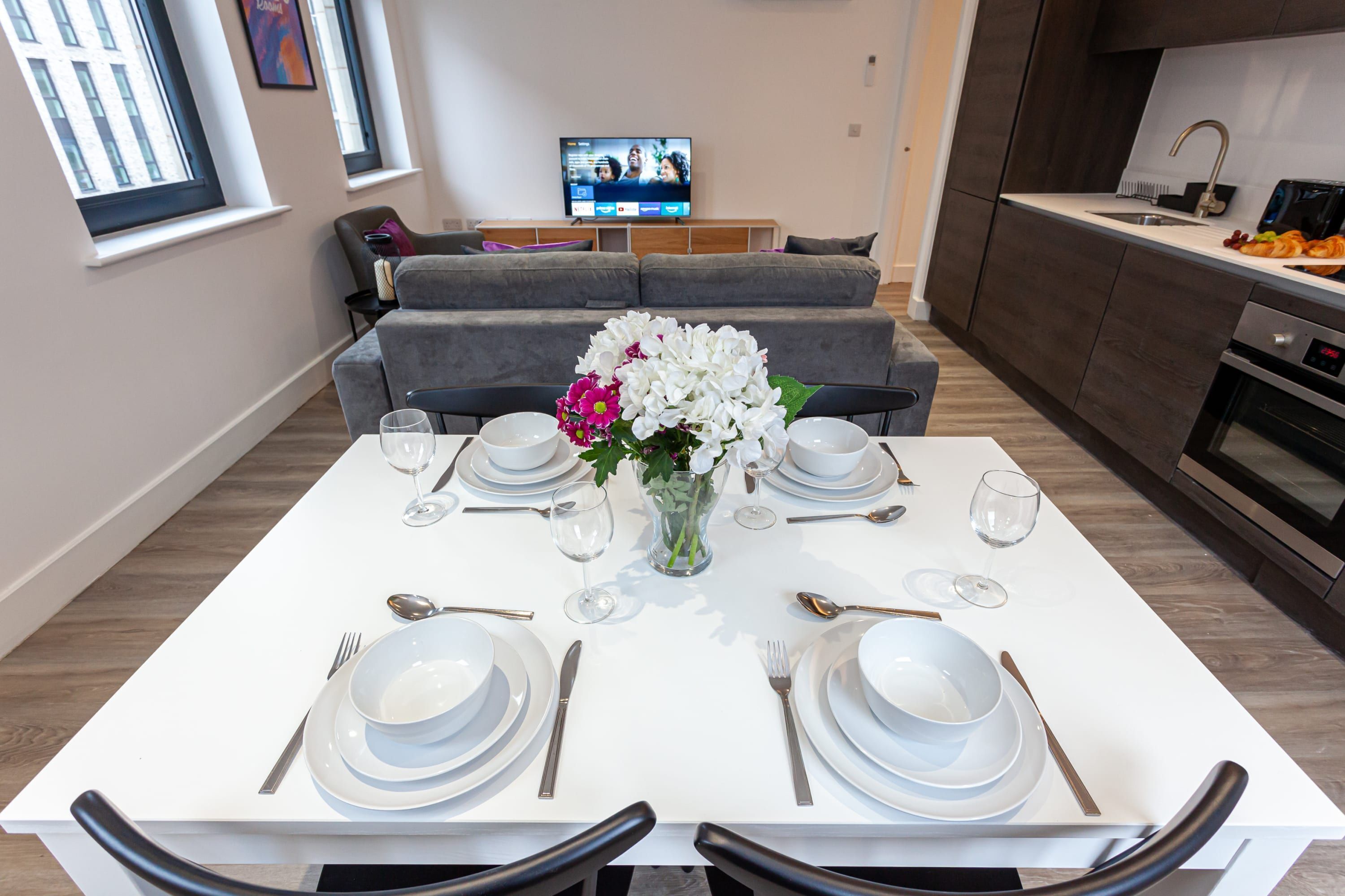 Uma sala de jantar com uma mesa branca posta para dois, um vaso com flores e uma TV ao fundo.