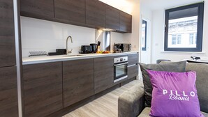 Una cucina moderna con mobili in legno scuro, una cappa in acciaio inossidabile e un forno integrato. Un cuscino viola con la scritta "PILLO Rooms" è su un divano grigio.