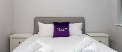 Un lit bien fait, avec deux serviettes blanches, un oreiller violet orné du texte « PILLO Rooms » et une tête de lit grise.
