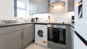 A modern kitchen with grey cabinets, a built-in washing machine, an oven, and a window with curtains. - Lovely One-bedroom Serviced Apartment in Eccles (Manchester)