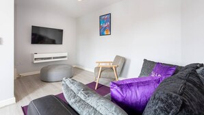 A modern living room with a grey sofa, a purple cushion, a wooden chair, a white entertainment unit, and a flat-screen TV. - Lovely One-bedroom Serviced Apartment in Eccles (Manchester)