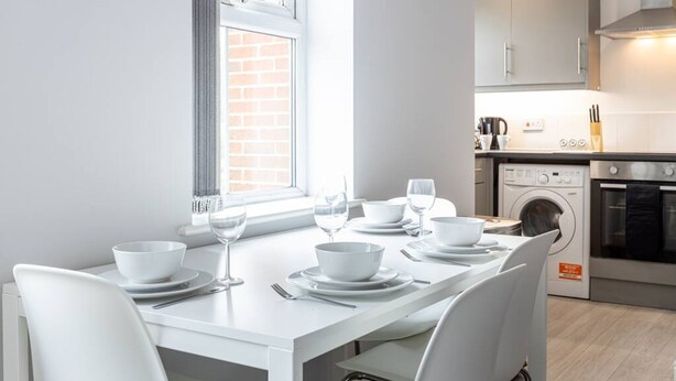 A modern kitchen with white cabinetry, a dining table set for four, and a washing machine in the background. - Lovely One-bedroom Serviced Apartment in Eccles (Manchester)