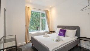 A neatly made bed with a grey headboard, a window with curtains, and a small bedside table.