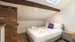 Un dormitorio con techo inclinado, vigas de madera a la vista, pared de ladrillo, tragaluz, cama con ropa blanca, mesita de noche y suelo de madera.