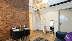 Un interior moderno con pared de ladrillo, una escalera, un comedor con mesa dispuesta para cuatro personas y un sofá con un cojín morado.