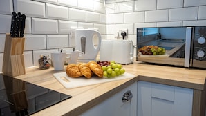 Una cocina moderna con baldosas blancas, un tostador, una tetera, una tabla de cortar con cruasanes y uvas, y un bloque de cuchillos.