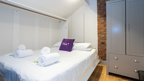 Una cama bien hecha con toallas enrolladas y una almohada morada, un armario gris y una pared de ladrillo.