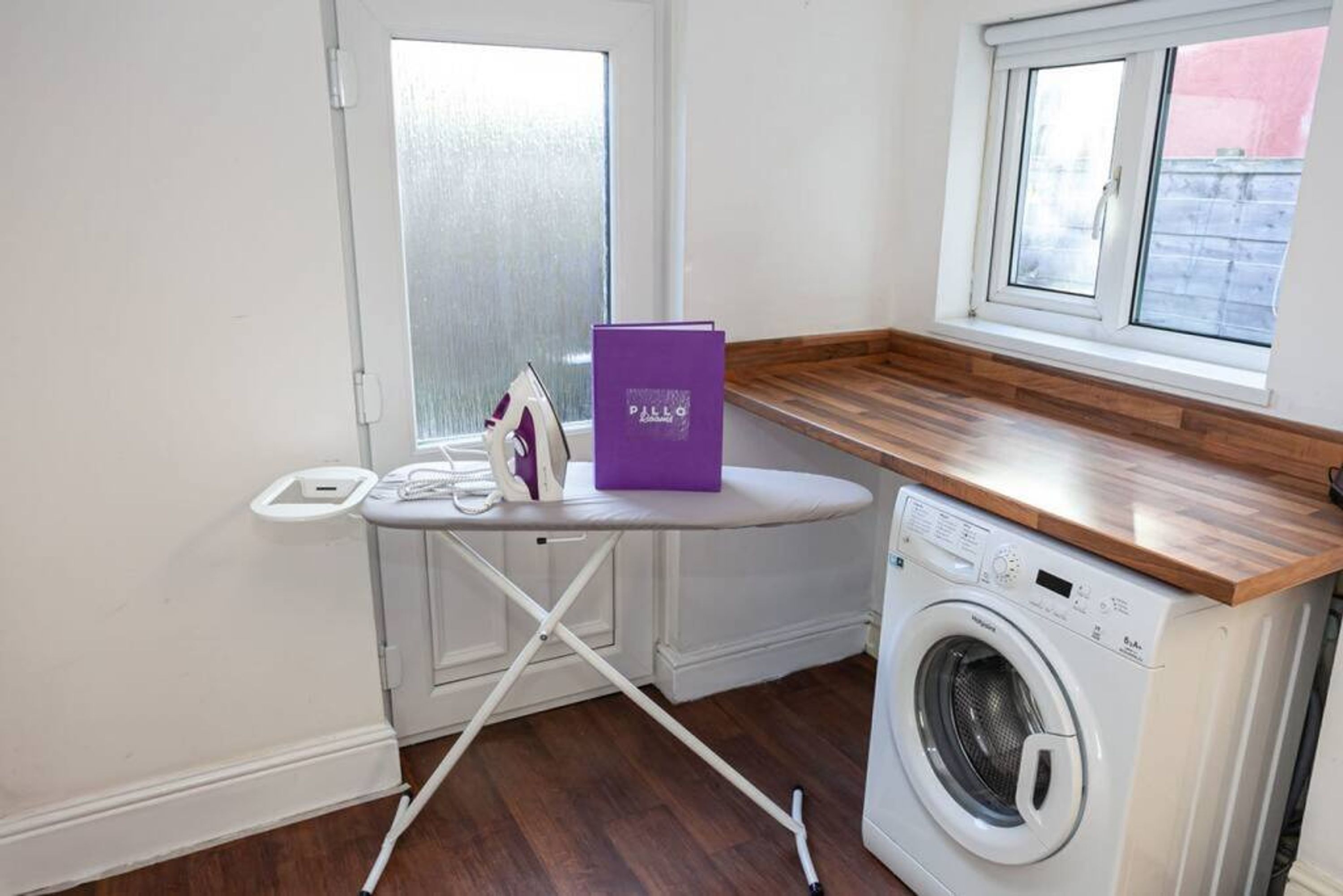 Un espace dédié à la lessive, comprenant un lave-linge, une planche à repasser et une boîte violette.