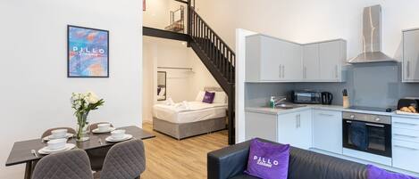 A modern apartment with a dining area, a living room with a black leather sofa, a kitchenette, and a staircase leading to a bedroom.