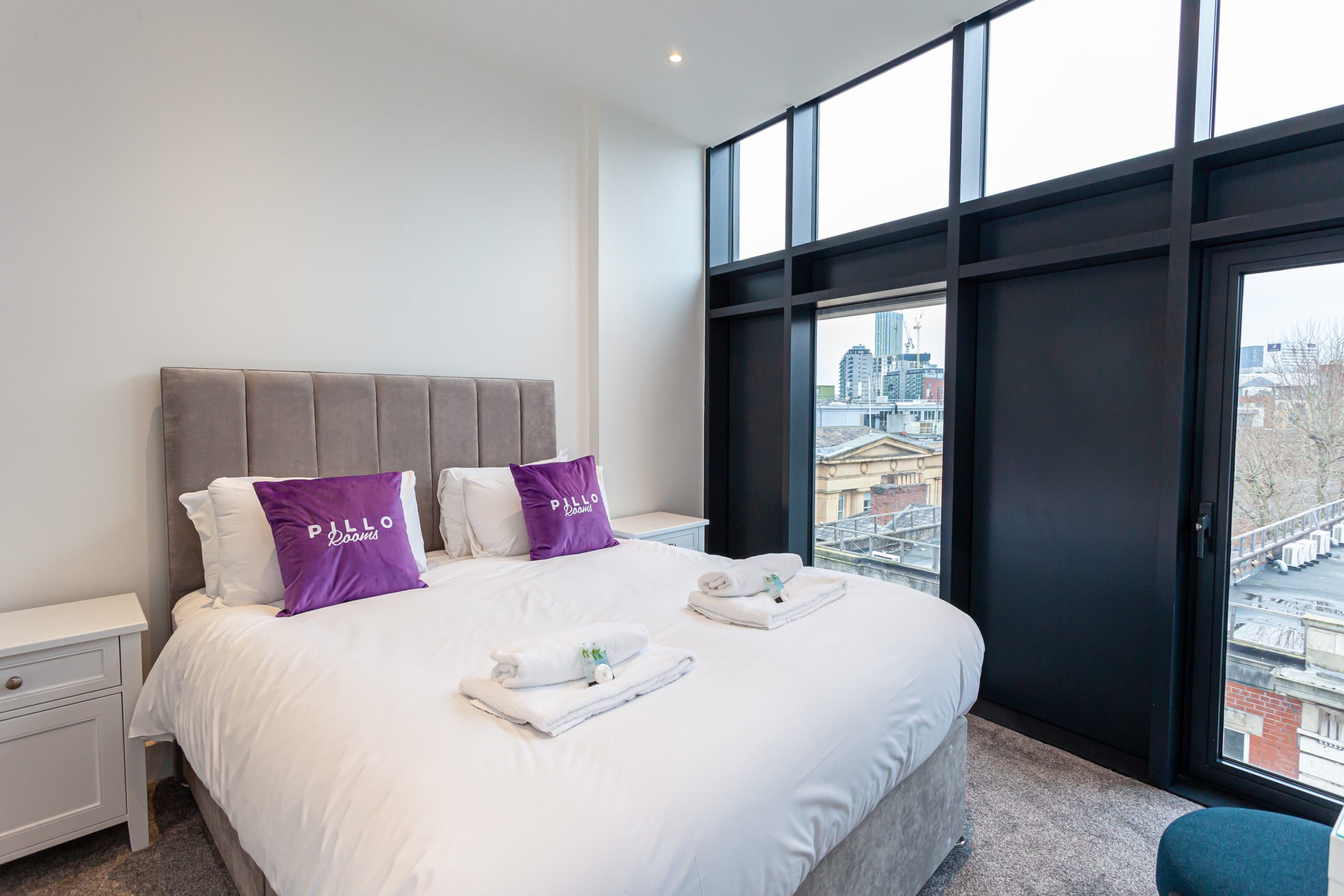 A modern hotel room with a large bed, two pillows, a nightstand, and a view of the city through large windows.