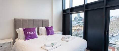 A modern hotel room with a large bed, two pillows, a nightstand, and a view of the city through large windows.