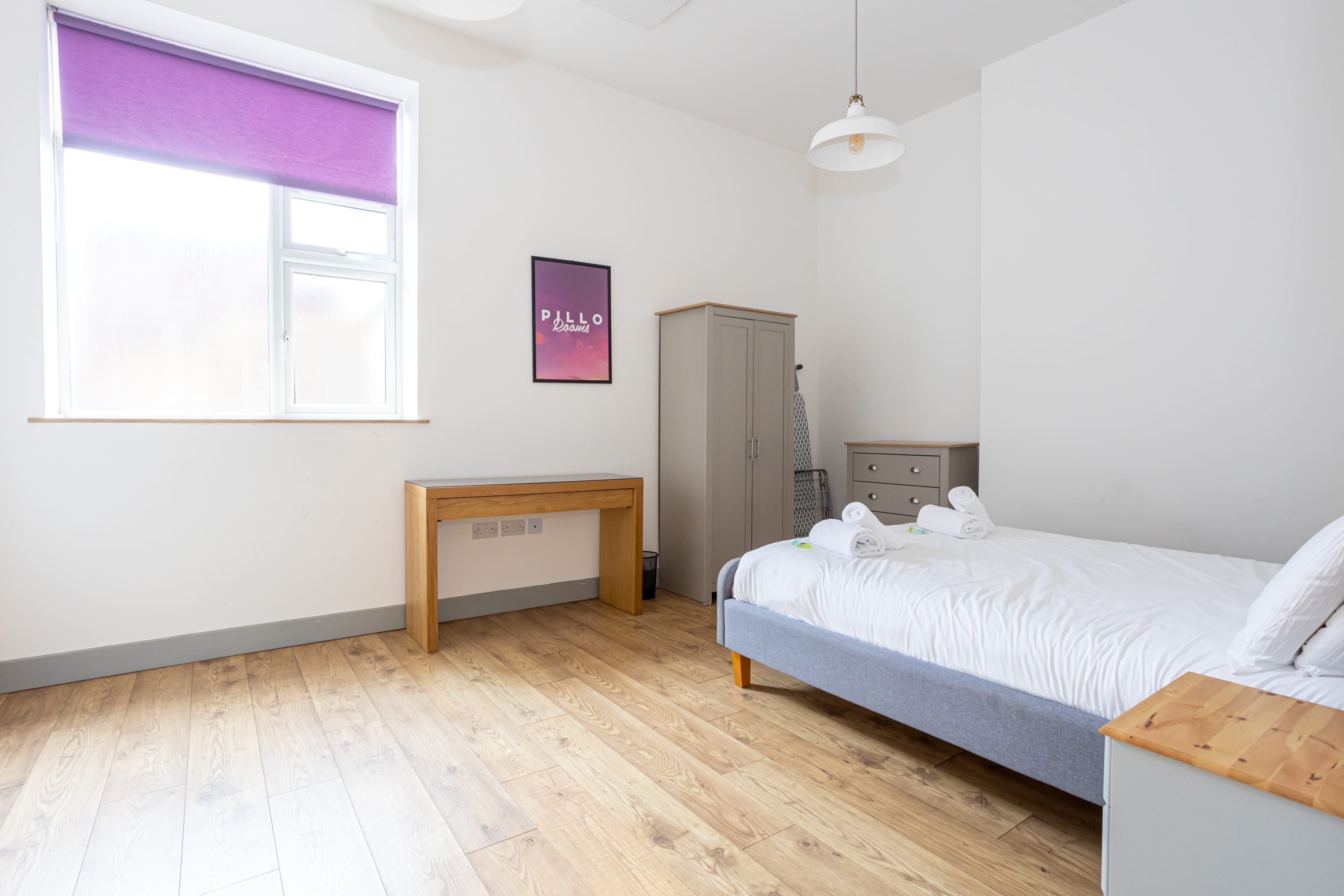 Une chambre à coucher comprenant un lit, un bureau, une armoire et une fenêtre avec un store violet.