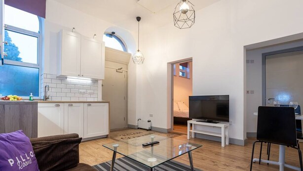 A modern living room with a glass coffee table, a sofa, and a TV. There is a kitchen with white cabinets and a window. - Unique 1 Bedroom Apartment by Pillo Rooms (Salford)