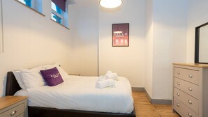 A neatly made bed with white linens and a purple pillow, a wooden nightstand, a dresser with drawers, and a framed sign on the wall. - Unique 1 Bedroom Apartment by Pillo Rooms (Salford)