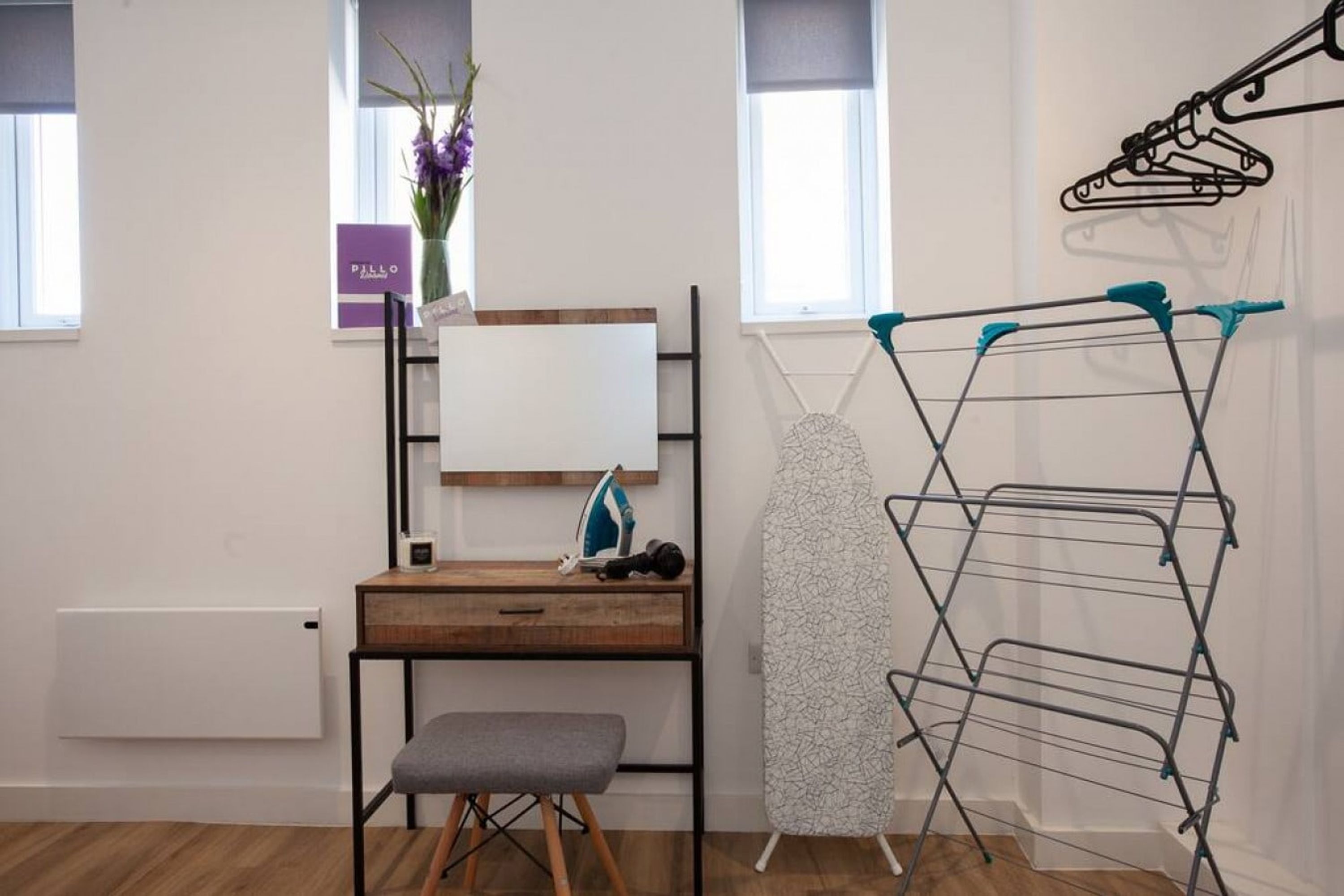 A room with a wooden desk, a grey bench, a wall-mounted ironing board, and a drying rack.