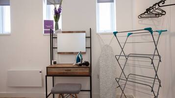 A room with a wooden desk, a grey bench, a wall-mounted ironing board, and a drying rack.