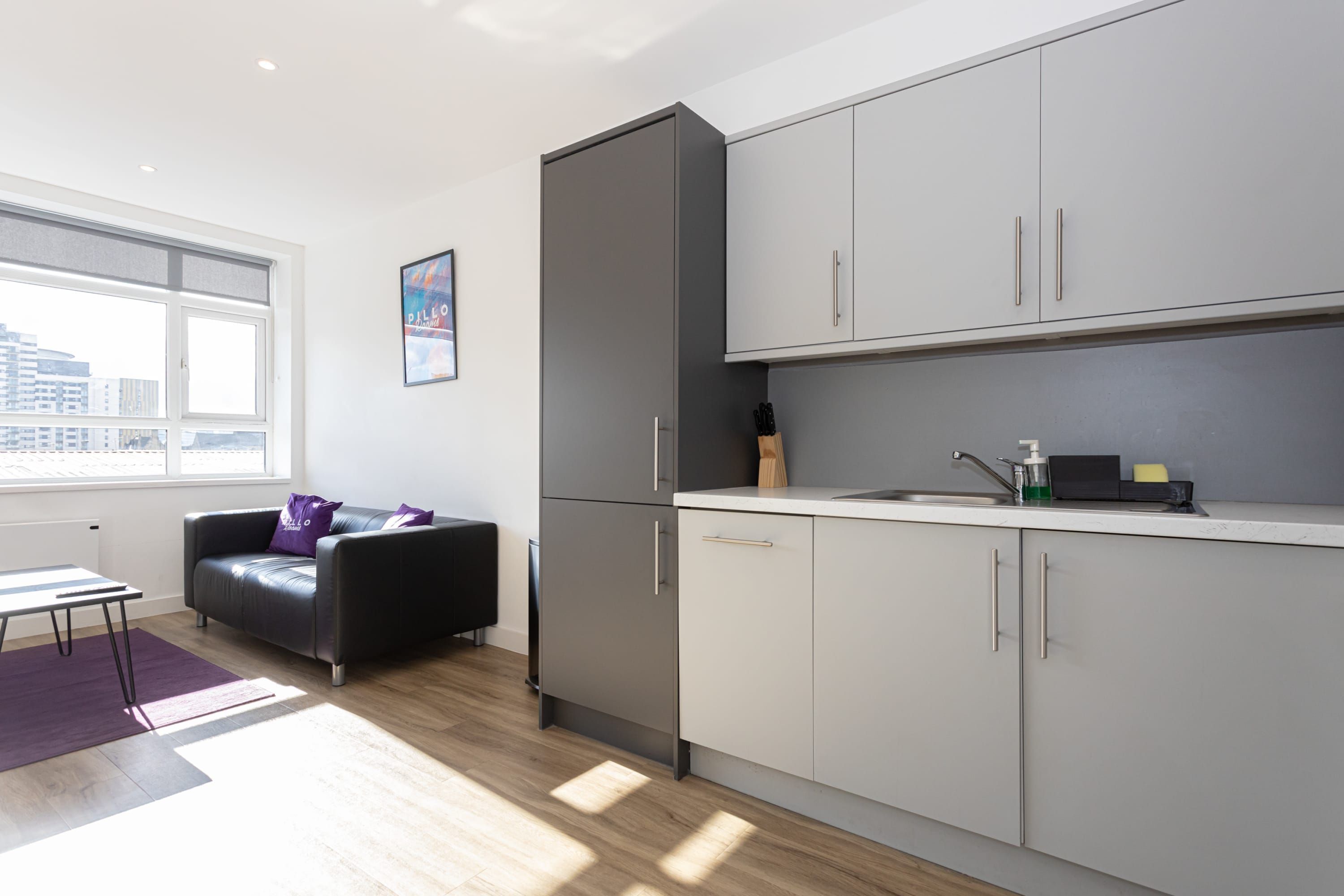 A modern kitchen with grey cabinets, a black sofa, and a window providing a view of the city.