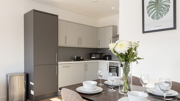 A modern kitchen with grey cabinets, a dining table set with white plates and glasses, and a vase with white flowers. - Modern, Stylish 1BR Apartment Nearby AO Arena (Manchester)