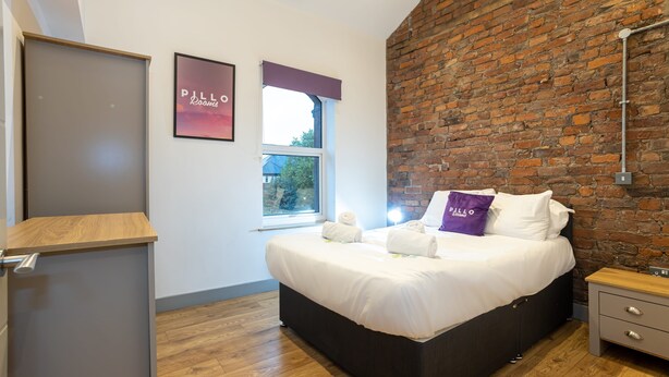 Un dormitorio con cama, mesita de noche, ventana con vistas, pared de ladrillo y puerta.