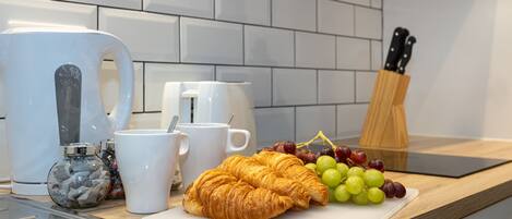 A kitchen counter with a coffee maker, a white mug, a cutting board with grapes and a croissant, and a knife block.