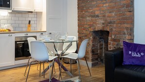 Una cocina moderna con armarios blancos, una pared de ladrillo y una zona de comedor con mesa de vidrio y sillas blancas.